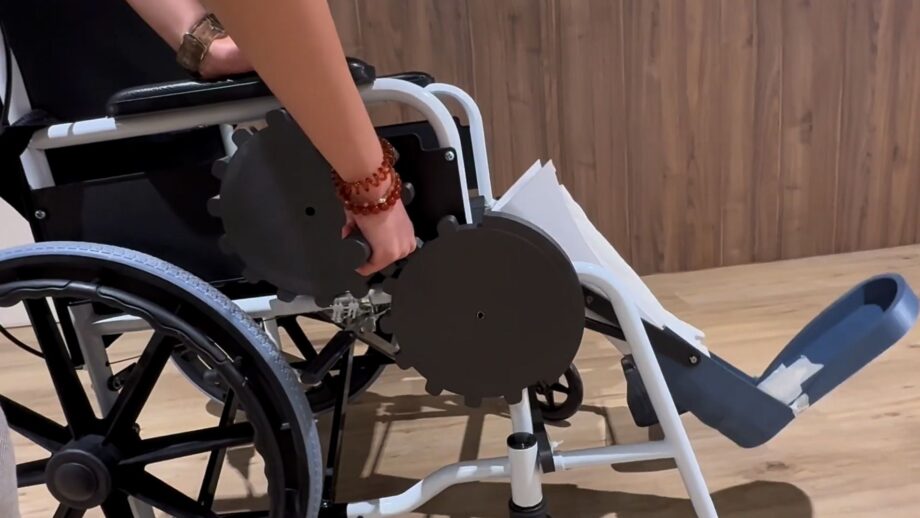 A wheelchair fitted with a custom leg elevation extension featuring mechanical gears and a lever, designed to help the user lift and support her leg comfortably while seated.