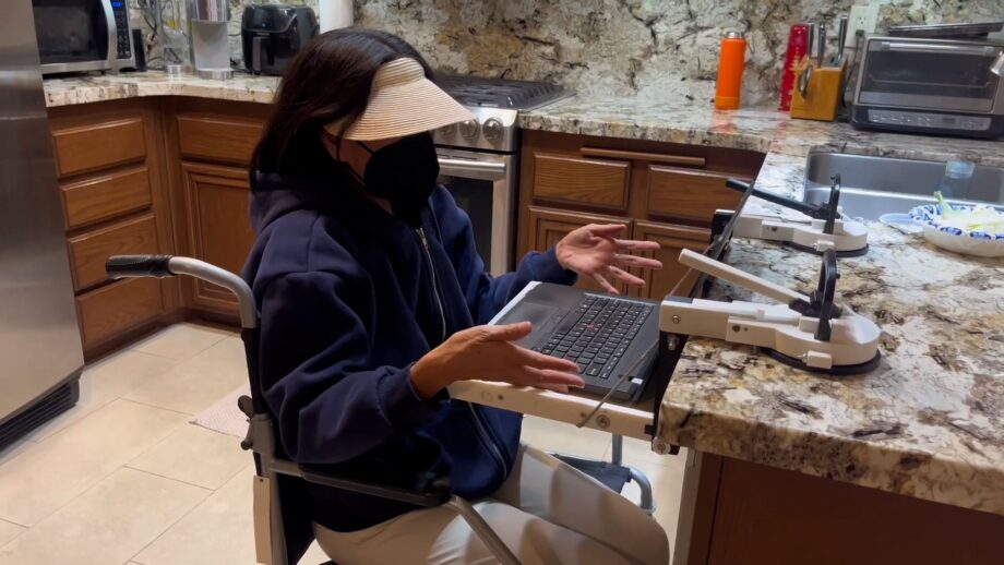 A person in a wheelchair using a laptop on a portable table that is attached to a kitchen unit.