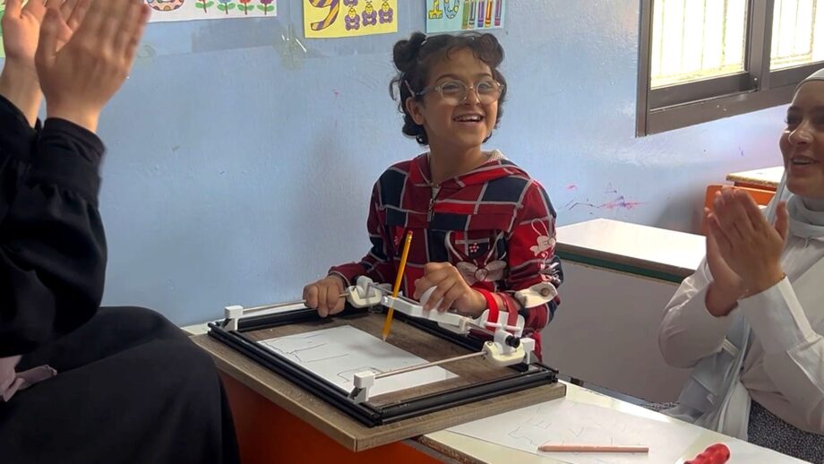 A young girl with hemiplegia smiles as she uses a rail-guided writing aid that supports her hand and helps her move a pen smoothly across the page.