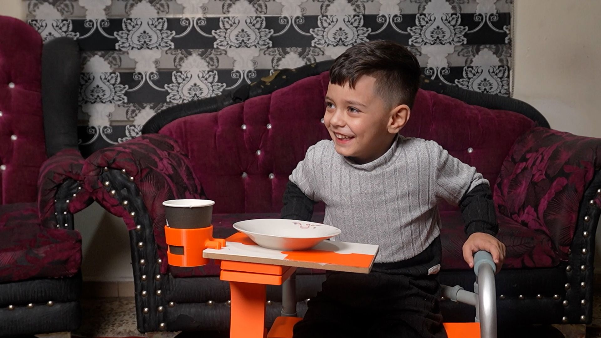 Young boy sitting beside a walker with a 3D printed orange tray and cup holder attached.